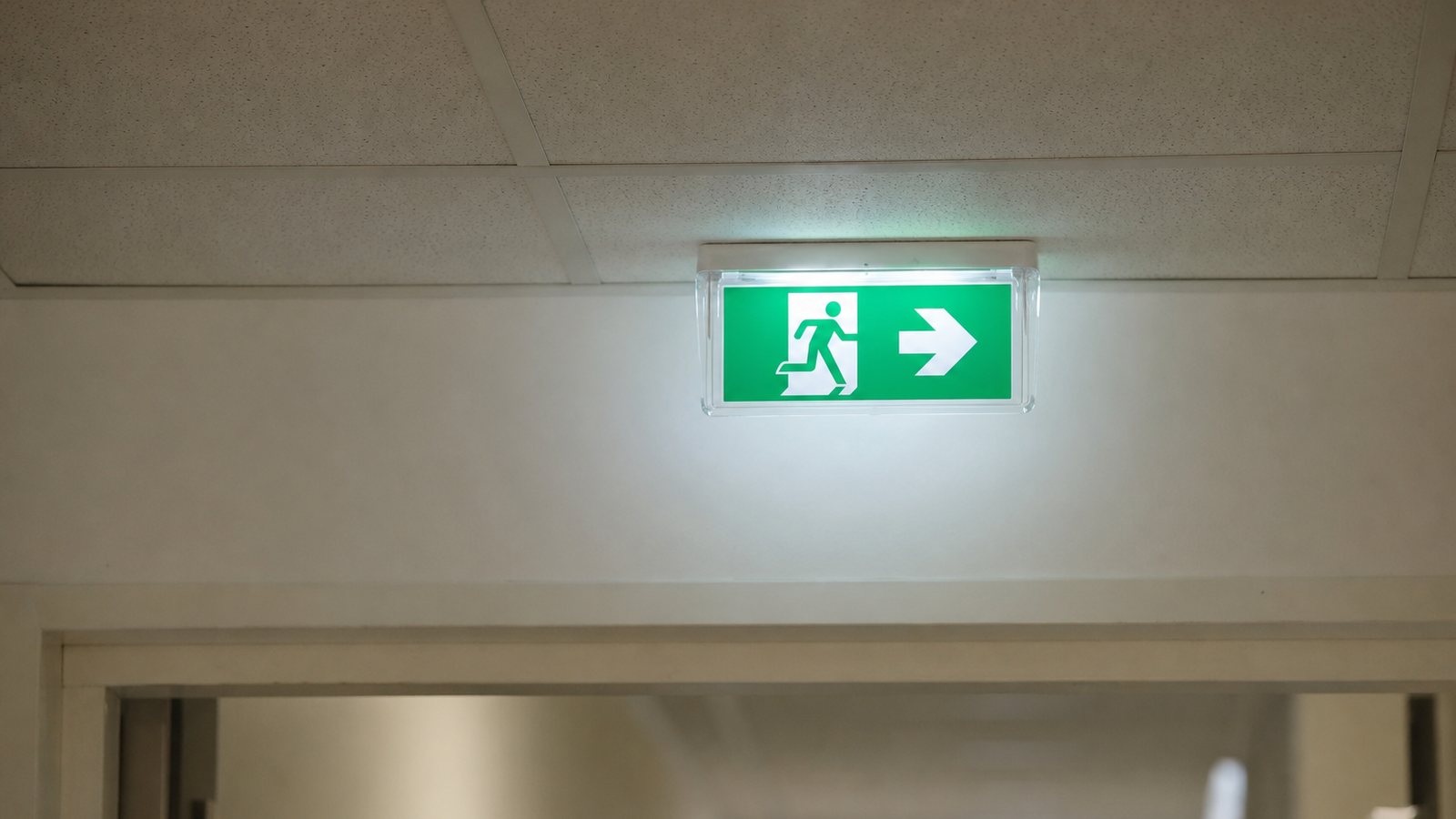 Bloc autonome d'éclairage de sécurité vert allumé au-dessus d'une porte de sortie dans un couloir